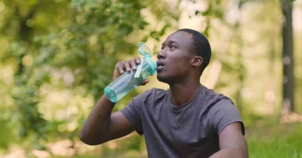Afrykański Amerykanin pijący wodę, odpoczywający w parku po treningu - Materiał filmowy, wideo
