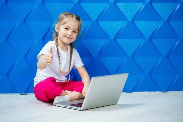 7 años de edad, niña en una camiseta blanca se sienta en el suelo con un ordenador portátil y presiona las teclas - Foto, imagen