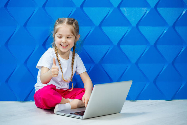 7-letnia dziewczyna w białej koszulce siedzi na podłodze z laptopem i naciska klawisze - Zdjęcie, obraz