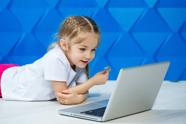 A modern child girl uses a laptop lying on the floor with legs looking at the camera. Funny kid on a bright blue wall background. Modern internet wireless technology - Foto, Imagen