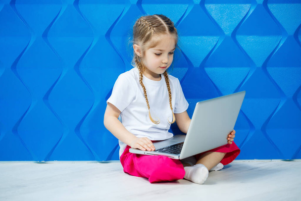 Moderní holčička používá notebook ležící na podlaze s nohama upřenýma do kamery. Vtipný kluk na jasně modrém pozadí. Moderní bezdrátová technologie internetu - Fotografie, Obrázek