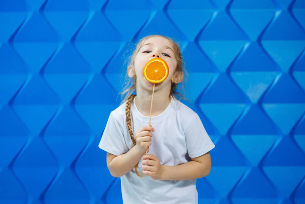klein gelukkig meisje met een oranje in haar hand, kijkt naar de camera gekleed in wit T-shirt, geïsoleerd op blauwe achtergrond, kopieer ruimte, - Foto, afbeelding
