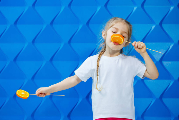Schattig meisje met sinaasappels ze eet ze op een blauwe achtergrond - Foto, afbeelding