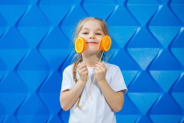 Cute girl z pomarańczy zjada je na niebieskim tle - Zdjęcie, obraz