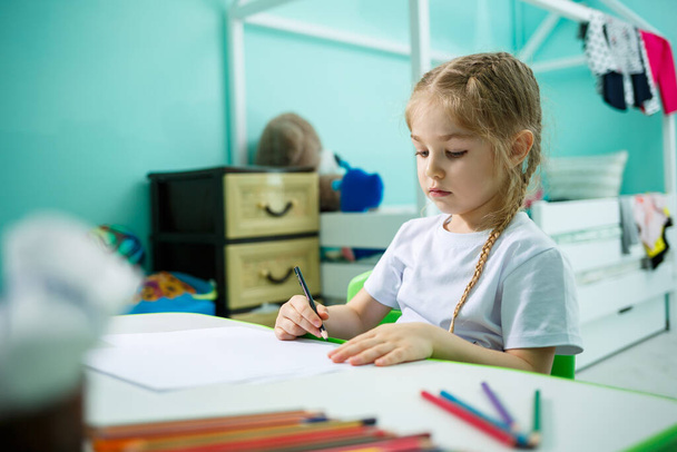Uma menina em uma camiseta branca senta-se em seu quarto na mesa e desenha com lápis de cor
 - Foto, Imagem