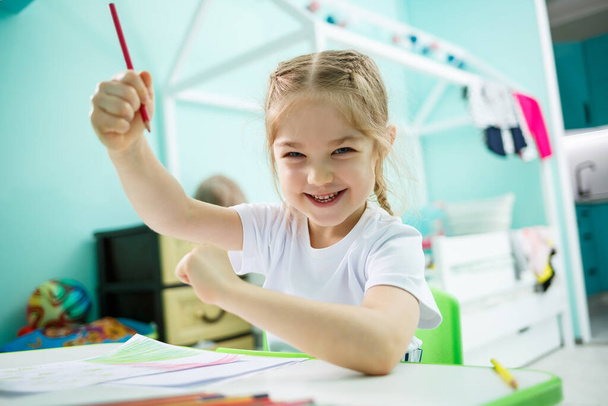 Urocza dziewczynka rysująca ołówkami w domu, siedząca przy stole. Kreatywne dziecko siedzące w pokoju i uczące się rysować. Maluch dziewczyna robi pracę domową w domu. - Zdjęcie, obraz