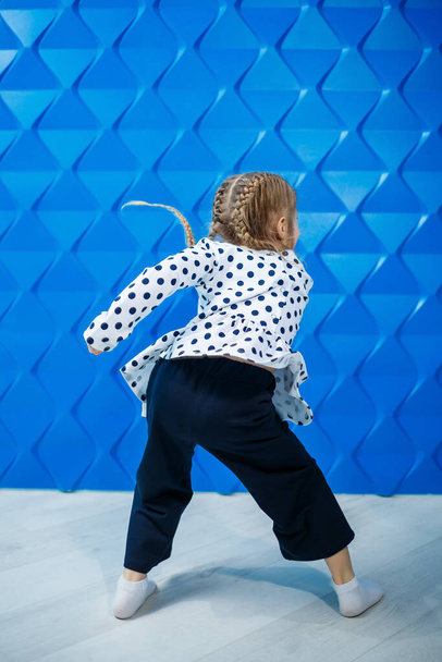 Blondynka 7-letnia w kurtce z groszkiem na niebieskim tle tańczy i śmieje się, radosne dziecięce emocje, szczęśliwe dzieciństwo - Zdjęcie, obraz