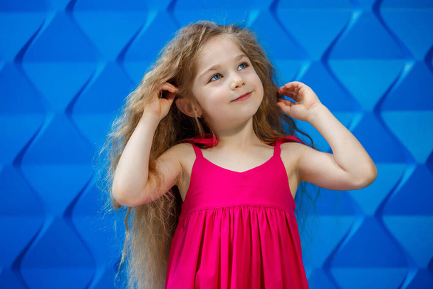 Ragazzina dai capelli biondi con un vestito rosa su uno sfondo blu che balla e ride, emozioni di gioia per bambini brillanti, infanzia felice - Foto, immagini