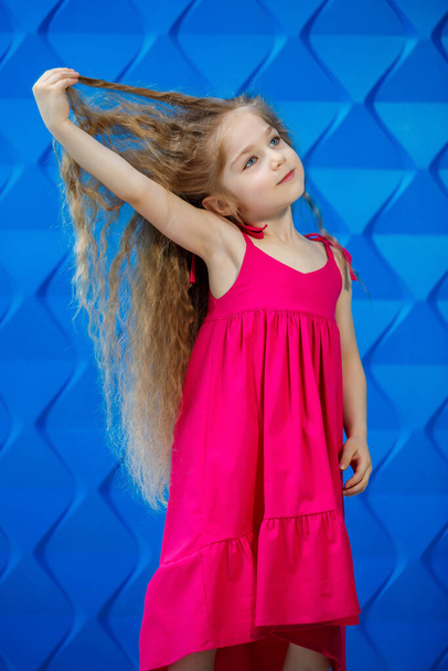 Szép hajú kislány rózsaszín ruhában, kék háttér-tánccal és nevetéssel, ragyogó gyerekek öröme, boldog gyermekkor - Fotó, kép