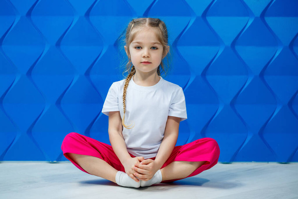 Menina com tranças em uma camiseta branca em um fundo azul. Ela se senta em uma posição de lótus no chão e sorri e acena suas mãos
. - Foto, Imagem