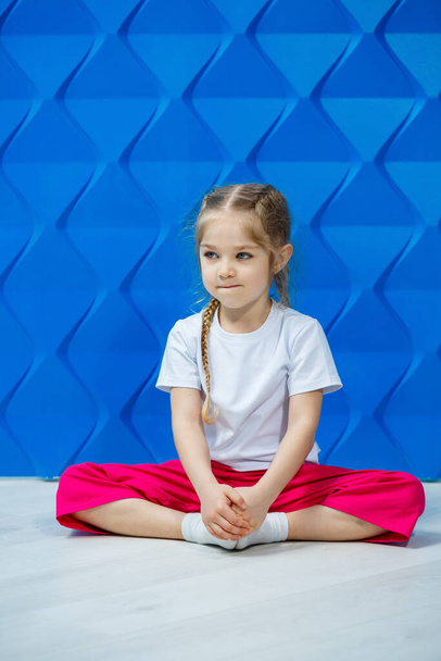 Little girl with pigtails in a white T-shirt on a blue background. She sits in a lotus position on the floor and smiles and waves her hands. - Photo, Image
