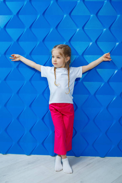 A girl with blond hair in a white T-shirt on a blue background. She smiles and waves her hands - Foto, Bild