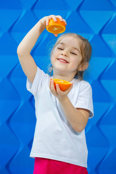 Pretty beautiful blonde girl in a white T-shirt smiles on a blue background. Fashionable cute child holds half an orange in his hands. The concept of an emotional portrait. - Photo, Image