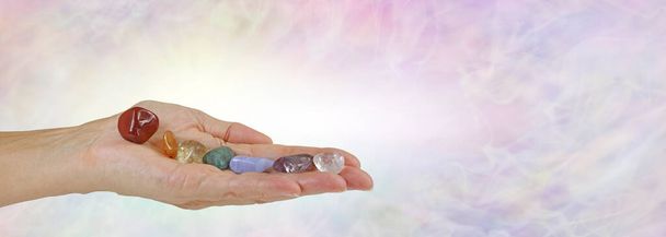 Seven Chakra Stones Mensagem Banner - mão feminina plana aberta com sete pedras caídas ordenadamente equilibradas em uma fileira contra um fundo de formação de energia wispy pálido com espaço de cópia - Foto, Imagem