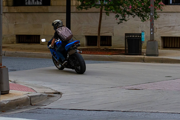 Un homme caucasien aux cheveux longs portant un bakcpack, un t-shirt, des gants, un jean et un casque conduit une moto de sport dans un endroit urbain. L'image le montre alors qu'il fait un virage à gauche à un carrefour. - Photo, image