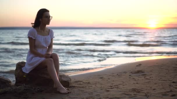 Egy fiatal nő ül egy kövön a tengerparton, fülhallgatóval a fülében. Egy fehér ruhás lány naplementekor zenét hallgat fejhallgatón.. - Felvétel, videó