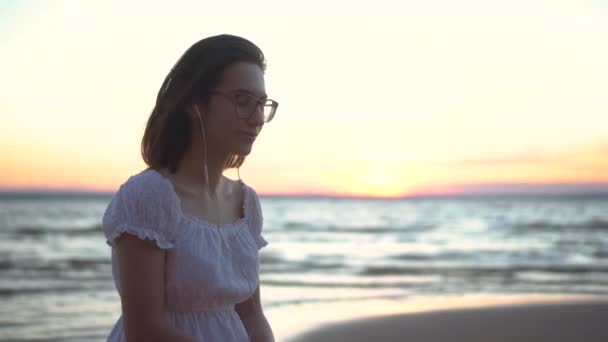 Une jeune femme est assise sur une pierre sur la plage au bord de la mer avec des écouteurs dans les oreilles. Une fille en robe blanche au coucher du soleil écoute de la musique sur écouteurs gros plan. - Séquence, vidéo