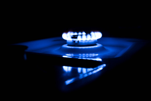 Llama azul de un quemador encendido en una estufa de gas. Llama iluminada sobre un fondo oscuro, foto tomada por la noche. Un ligero reflejo en la carcasa blanca de la estufa de gas. - Foto, imagen