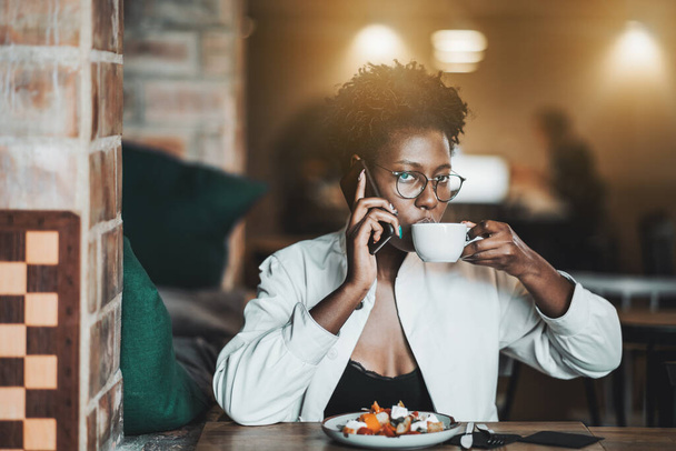 Uma jovem atraente e elegante mulher negra em óculos e uma capa branca está bebendo um delicioso café com leite e conversando ao telefone enquanto almoça em um café; uma mulher africana com café em um restaurante - Foto, Imagem