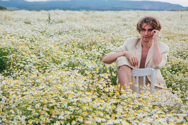 Tall handsome man dressed in a white suit on naked body sitting on a white chair in camomile flowers field - Zdjęcie, obraz