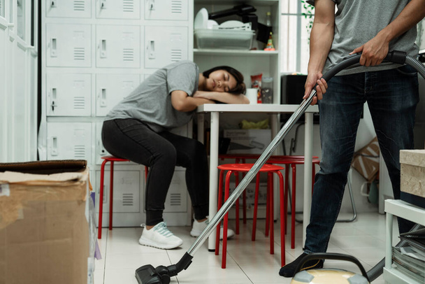 Un homme nettoie la pièce en utilisant l'aspirateur pendant que la fille dort sur la table. - Photo, image
