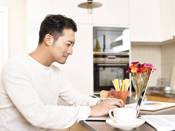 joven asiático hombre de negocios trabajando desde casa sentado en cocina contador usando laptop ordenador - Foto, imagen