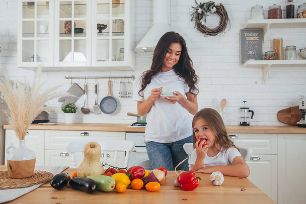 Szczęśliwa matka i dziecko pozują w domu. Piękna młoda mama i córeczka bawiące się i przygotowujące warzywa do sałatki w białej kuchni w skandynawskim stylu wnętrza. Zdrowa żywność - Zdjęcie, obraz