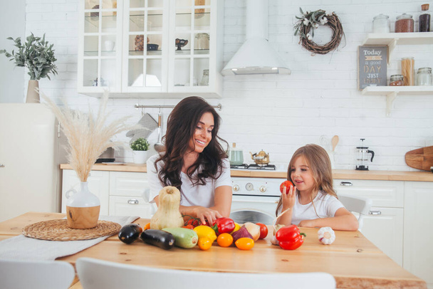 Gelukkige familie moeder en kind poseren thuis. Mooie jonge moeder en dochtertje die zich vermaken en groenten klaarmaken voor salade in een witte keuken in een Scandinavisch interieur. Gezond voedsel - Foto, afbeelding