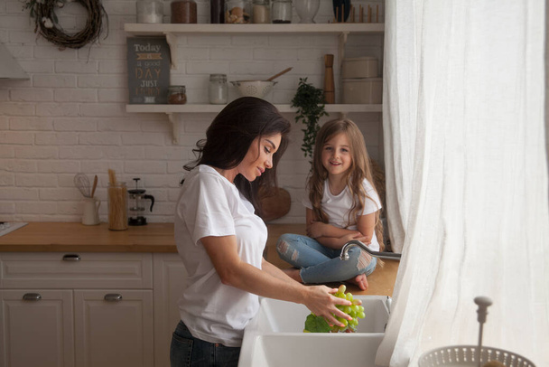 Feliz familia amorosa preparando la comida juntos. Sonriente Mamá y su hija niña lavando frutas y verduras y divirtiéndose en la cocina. Pequeño ayudante en el interior de estilo escandinavo. Alimento saludable - Foto, Imagen