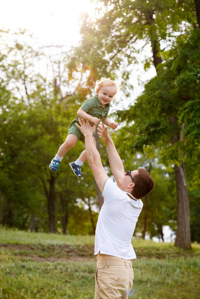 Junger Vater und kleiner Sohn. Papa wirft seinen Sohn gegen die Bäume im Hintergrund. Spaziergang im Park. Familienleben. Vertikal gerahmte Aufnahme. - Foto, Bild