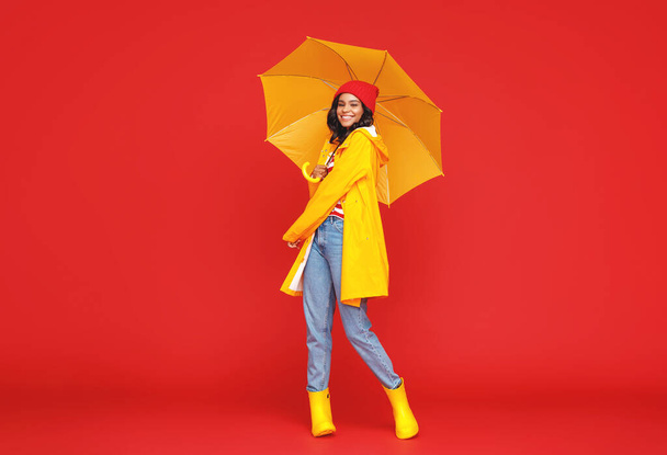 Plná délka optimistické etnické ženy s deštníkem s úsměvem a při pohledu do kamery na deštivý podzimní den proti červenému pozadí - Fotografie, Obrázek