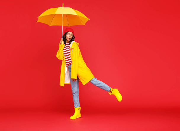 Full length cute ethnic woman with umbrella smiling and looking away on rainy autumn day against red backgroun - Zdjęcie, obraz