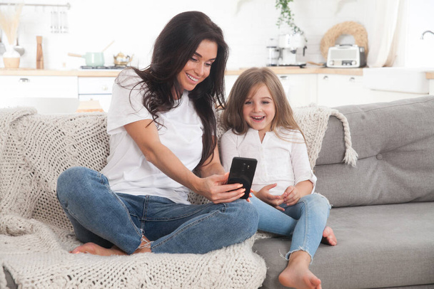 Attractive young mom and little daughter playing online games at home using smartphone, having video chat or watching movie. Distance education. Video conference or virtual event on quarantine - Foto, imagen