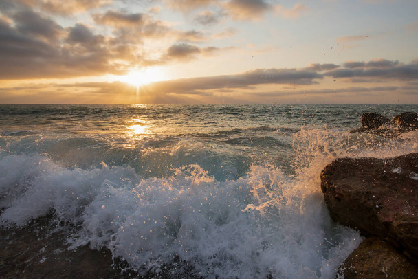 海の地平線に沈む太陽とともに、日没の海の波が海岸で砕けていっています - 写真・画像