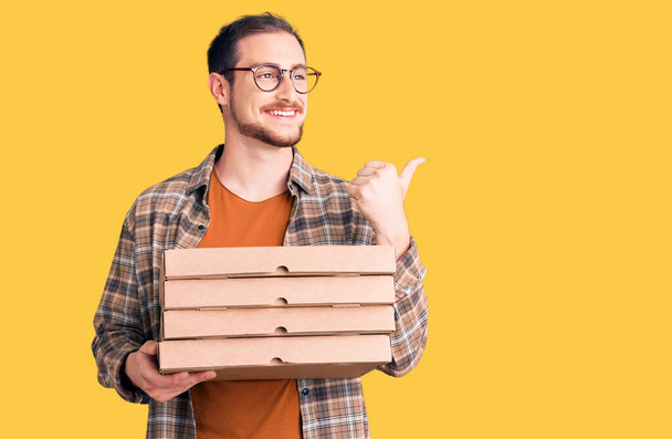Junge gutaussehende kaukasische Mann mit Lieferung Pizza-Box zeigt Daumen nach oben zur Seite lächelt glücklich mit offenem Mund  - Foto, Bild