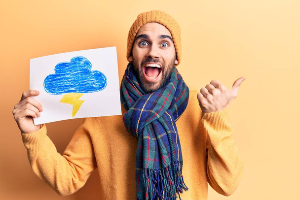 Νεαρός όμορφος άντρας με γενειάδα φορώντας χειμωνιάτικα ρούχα κρατώντας βροντές τραβώντας τον αντίχειρα στο πλάι χαμογελώντας χαρούμενος με ανοιχτό στόμα  - Φωτογραφία, εικόνα