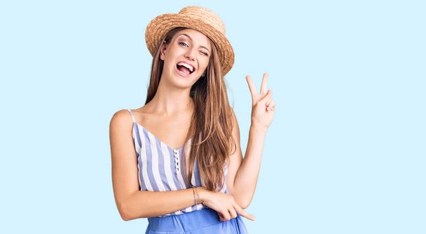 Young beautiful blonde woman wearing summer hat smiling with happy face winking at the camera doing victory sign with fingers. number two.  - Foto, Bild