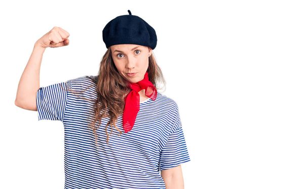 Jonge mooie blonde vrouw dragen franse baret en gestreept t-shirt sterke persoon tonen arm spier, vertrouwen en trots op de macht  - Foto, afbeelding