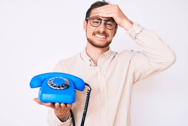 Mladý pohledný muž drží retro telefon vystresovaný a frustrovaný s rukou na hlavě, překvapený a rozzlobený obličej  - Fotografie, Obrázek