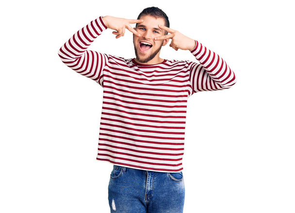 Young handsome man wearing striped sweater doing peace symbol with fingers over face, smiling cheerful showing victory  - Photo, Image