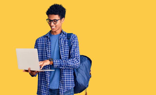 młody Afroamerykanin człowiek gospodarstwa student plecak za pomocą laptopa patrząc pozytywnie i szczęśliwy stojąc i uśmiechając się z pewnym uśmiechem pokazując zęby  - Zdjęcie, obraz