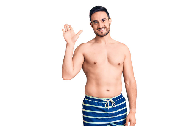 水泳ウェアを身に着けている若いハンサムな男はこんにちは幸せと笑顔、優しい歓迎ジェスチャーを言う  - 写真・画像