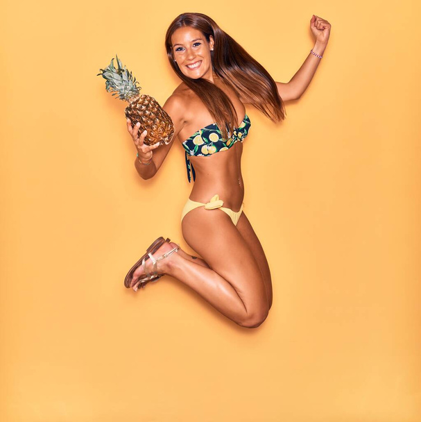Młoda, piękna Latynoska kobieta w bikini uśmiechnięta. Skoki z uśmiechem na twarzy trzymając ananasa na odizolowanym żółtym tle. - Zdjęcie, obraz