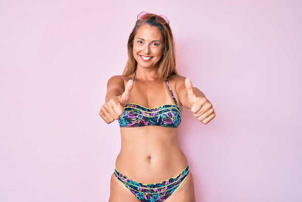Mujer hispana de mediana edad con bikini que aprueba hacer un gesto positivo con la mano, pulgares arriba sonriendo y feliz por el éxito. gesto ganador.  - Foto, imagen
