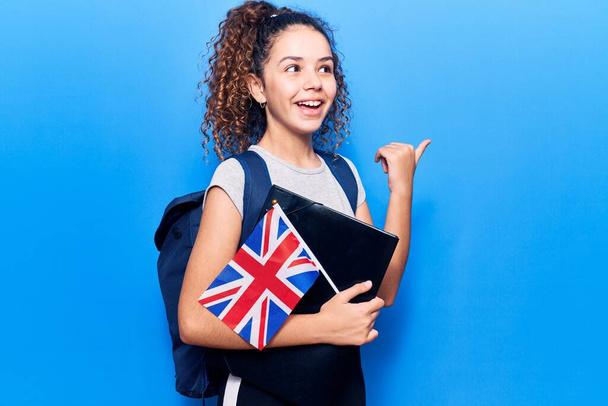 Gyönyörű gyerek lány göndör haj visel diák hátizsák gazdaság kötőanyag és Egyesült Királyság zászló mutató hüvelykujj fel az oldalra mosolyogva boldog nyitott szájjal  - Fotó, kép
