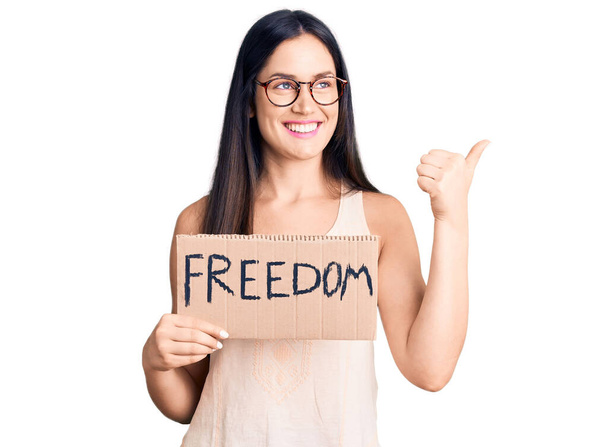 Молода красива біла жінка тримає прапор свободи, вказуючи великий палець в сторону посміхаючись щасливим з відкритим ротом
  - Фото, зображення