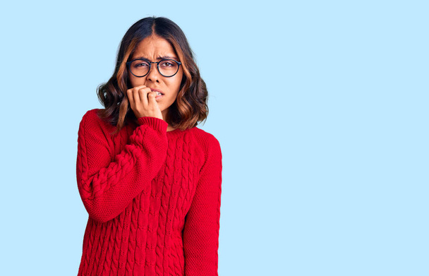 Young beautiful mixed race woman wearing red sweater and glasses looking stressed and nervous with hands on mouth biting nails. anxiety problem.  - Photo, Image