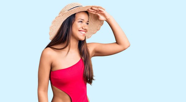 Joven chica latina hermosa con traje de baño y sombrero de verano muy feliz y sonriente mirando lejos con la mano sobre la cabeza. concepto de búsqueda.  - Foto, imagen