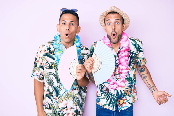 Jeune couple gay de deux hommes portant chemise d'été tenant fan de la main effrayé et étonné avec la bouche ouverte pour la surprise, visage incrédule  - Photo, image