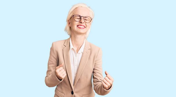 Young blonde woman wearing business clothes and glasses very happy and excited doing winner gesture with arms raised, smiling and screaming for success. celebration concept.  - Foto, afbeelding